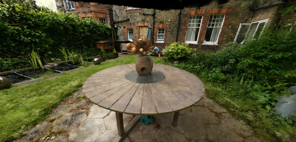 A Gaussian splat reconstruction of a garden, showing a round wooden table, planting, and a brick house behind it.